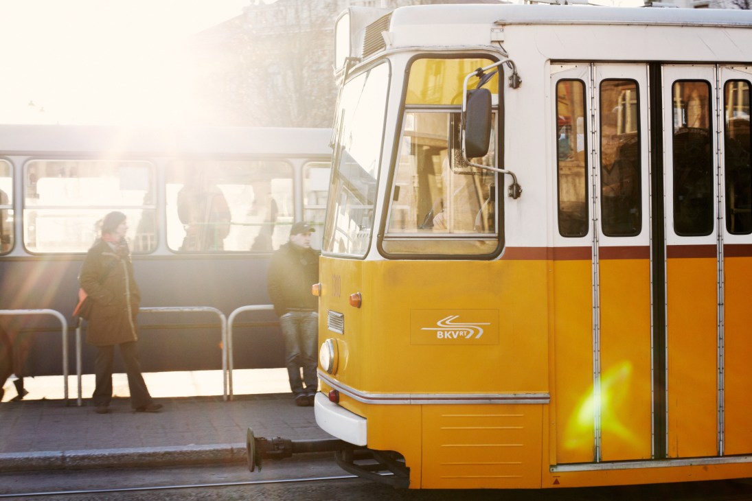 A yellow and white Budapest tram with passengers boarding at a street stop in morning sunlight.