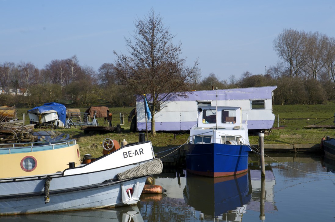 Moored boats and houseboats with green fields and bare trees in the background near Schellingerwoude.