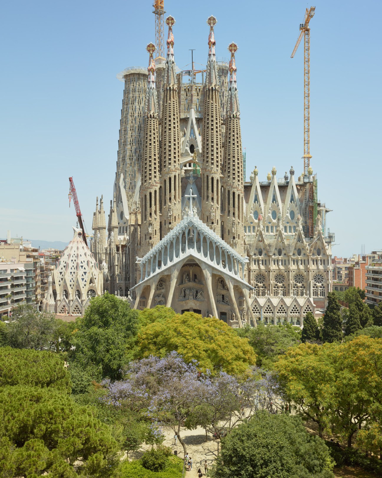 Sagrada Família basilica with multiple ornate spires, construction cranes, and surrounding Barcelona buildings and green trees