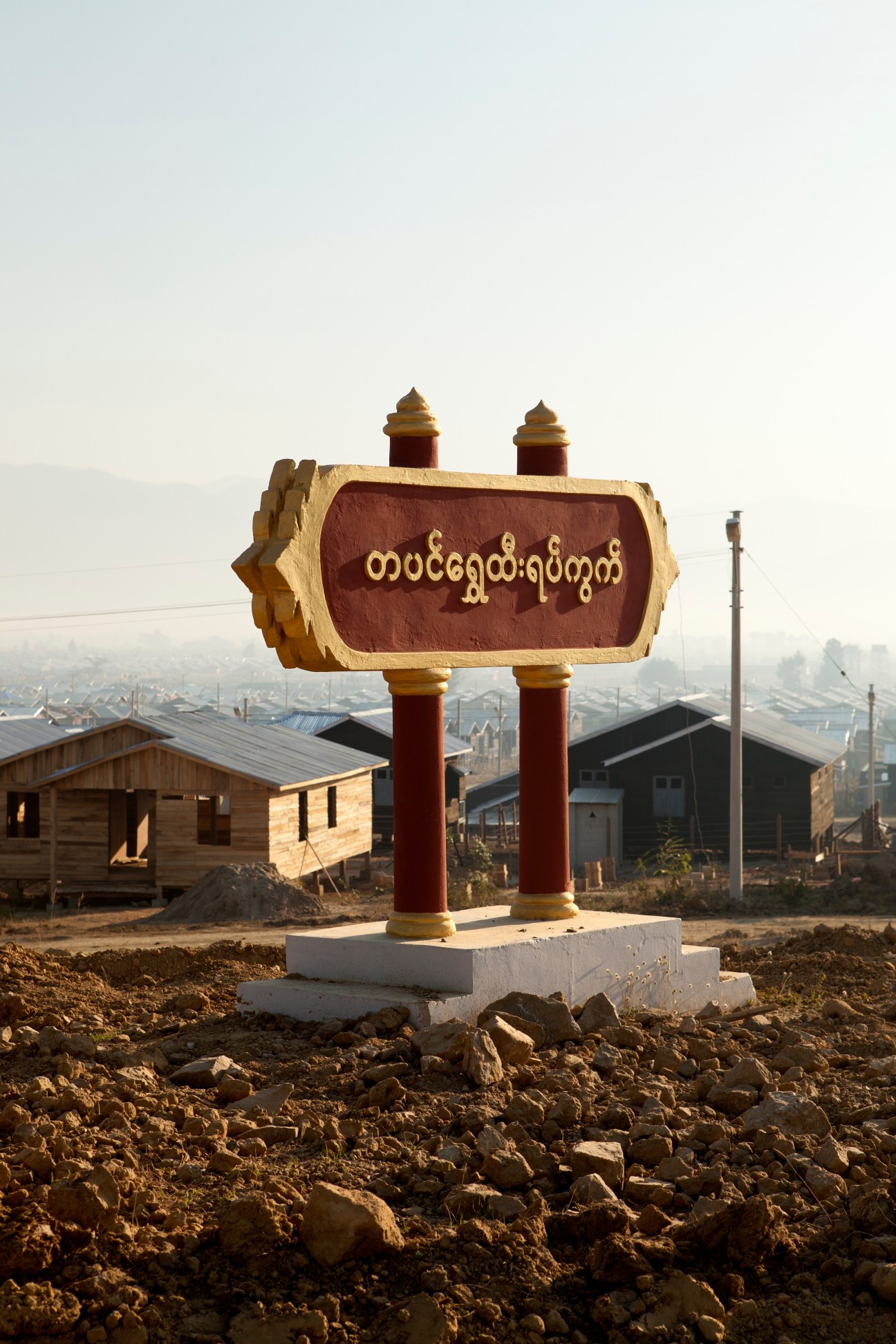 Ornate wooden sign with gold and red trim displaying Burmese script overlooking Popepa Thiri Town residential district in Naypyidaw.