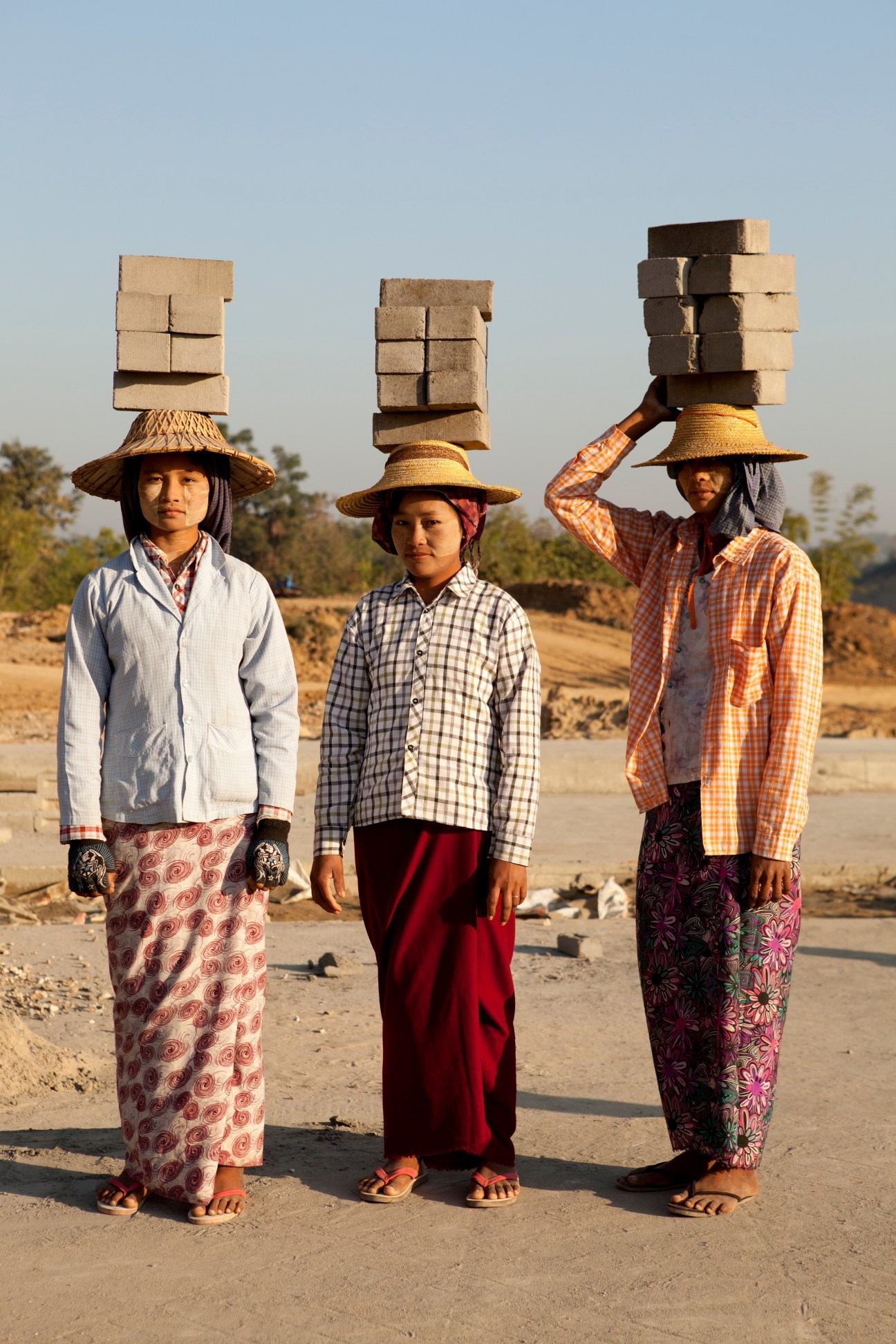 Three construction workers in traditional Burmese dress balance stacks of clay bricks on their heads.