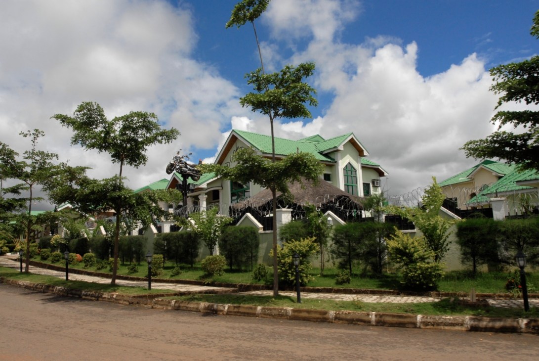 A white mansion with a green roof surrounded by manicured lawns and young trees in a gated residential development.