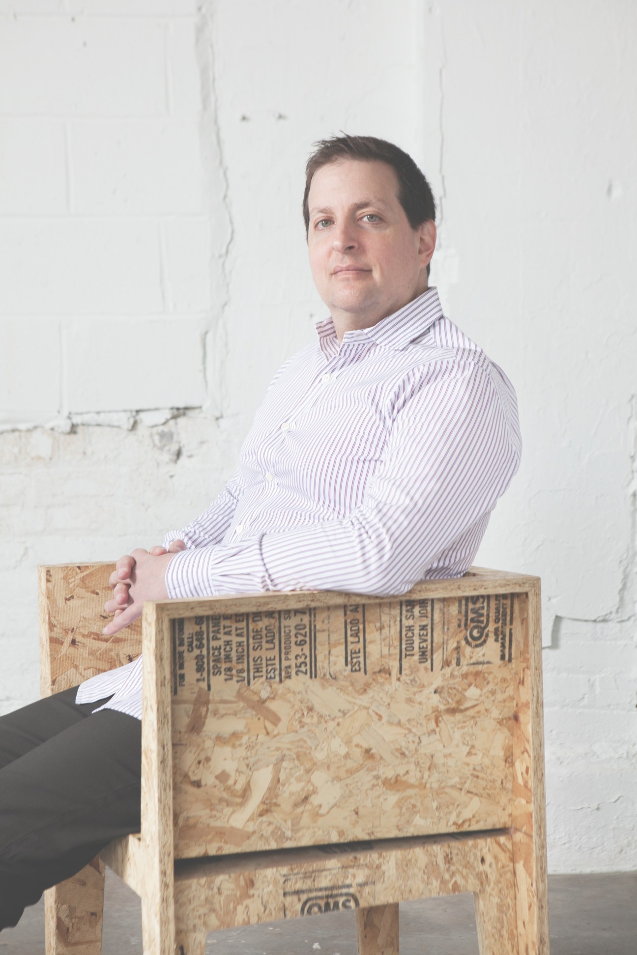 Man in purple striped shirt sitting on wooden crate against white wall