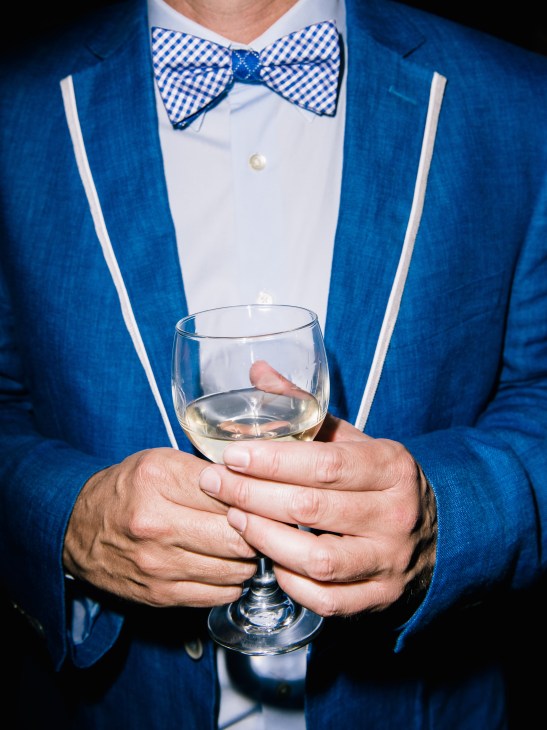 Man in blue blazer and checkered bow tie holding a glass of whiskey.