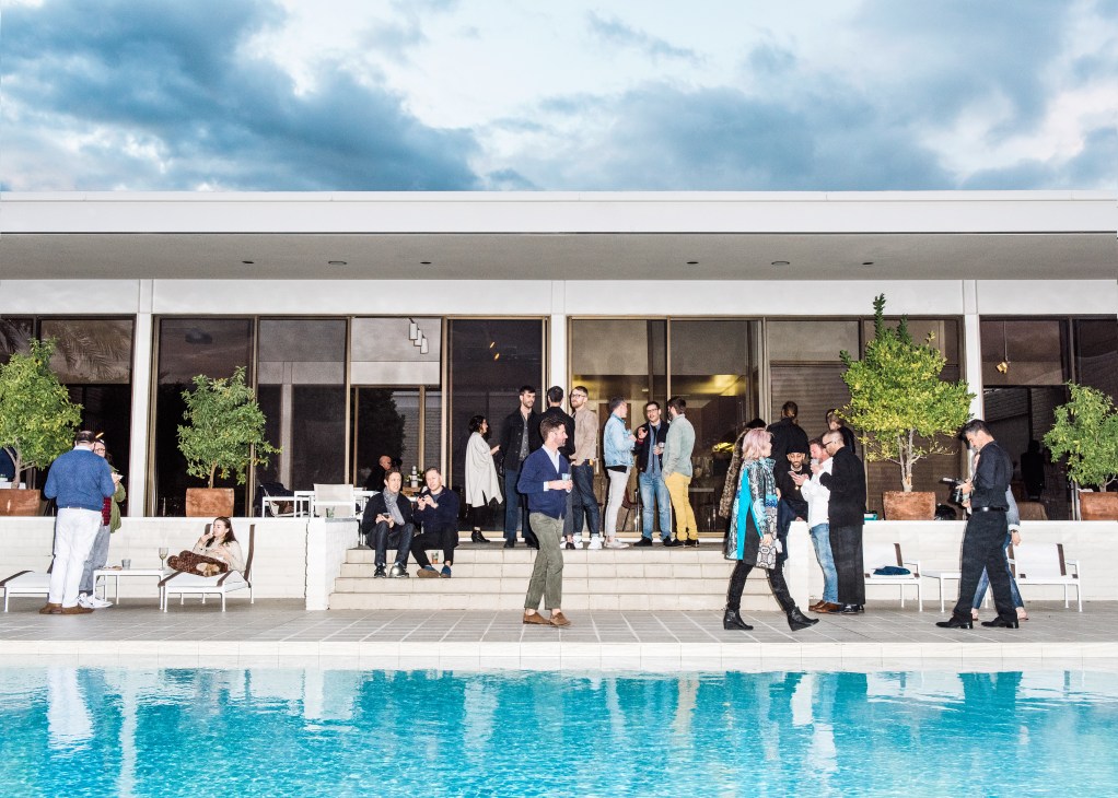 Guests mingling at a poolside cocktail party at a modernist mansion with expansive glass walls.