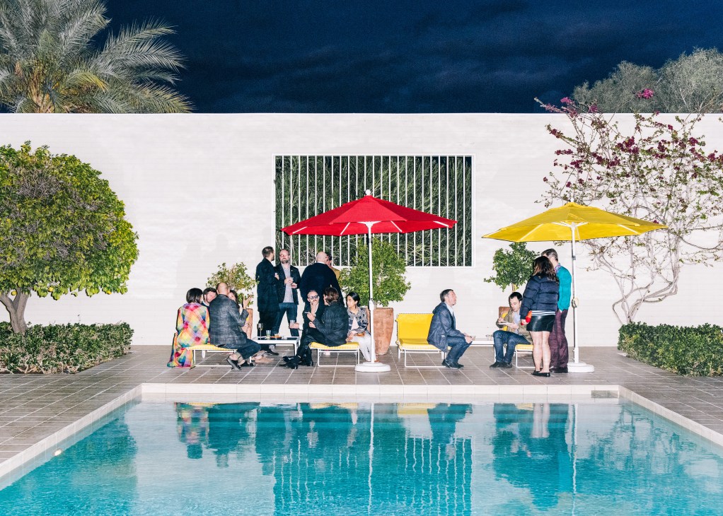 Guests gather by a modernist pool under red and yellow umbrellas at a mid-century Palm Springs residence.
