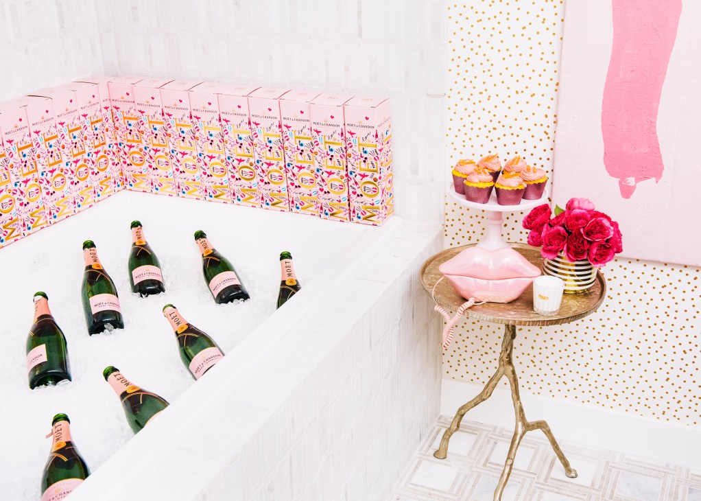 Styled flat-lay display featuring Moët & Chandon champagne bottles, cupcakes, pink lip-shaped dish, red roses, and gold-striped vase against patterned backdrop.