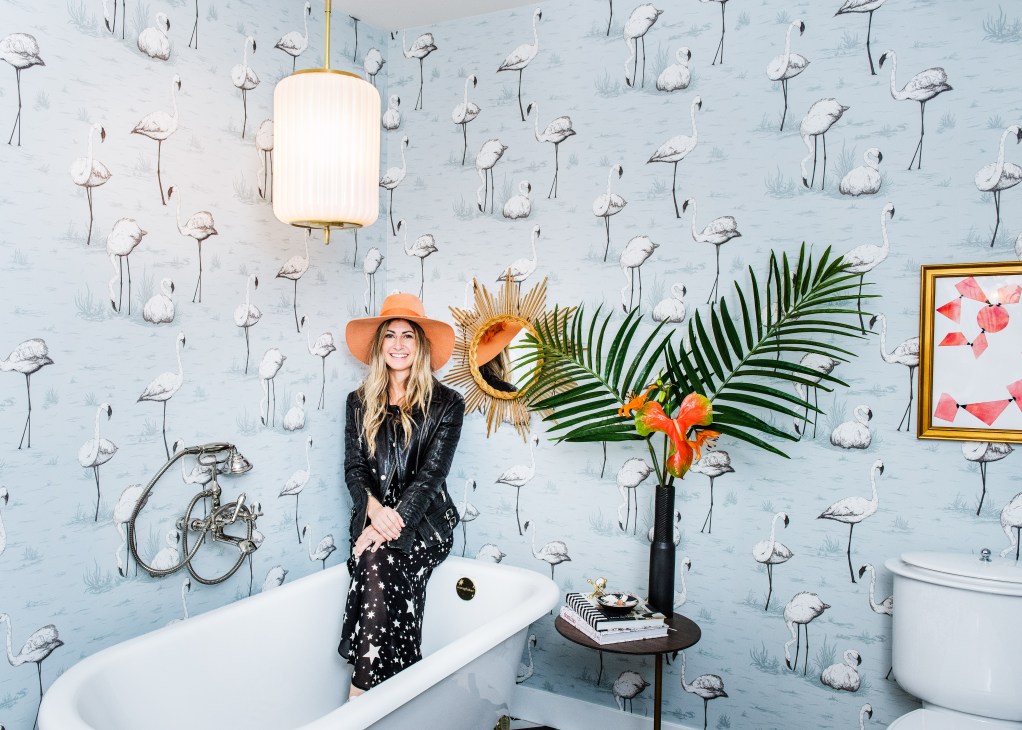 LA decorator Dee Murphy poses in a whimsical bathroom with flamingo-patterned wallpaper and mid-century modern fixtures.