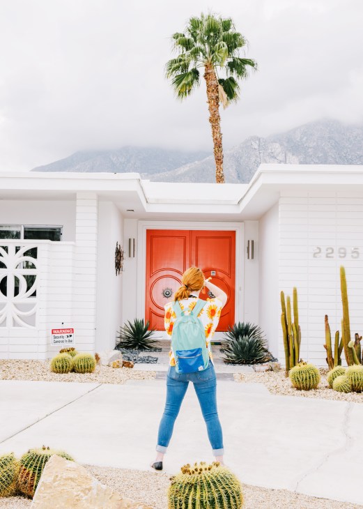 Woman standing on driveway facing a mid-century modern white house with bright red double doors and desert landscaping.