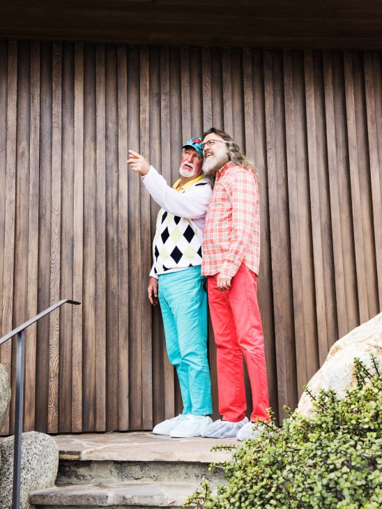 Two men standing against a wooden-slatted wall at the Edris House, one pointing while wearing colorful golf attire.