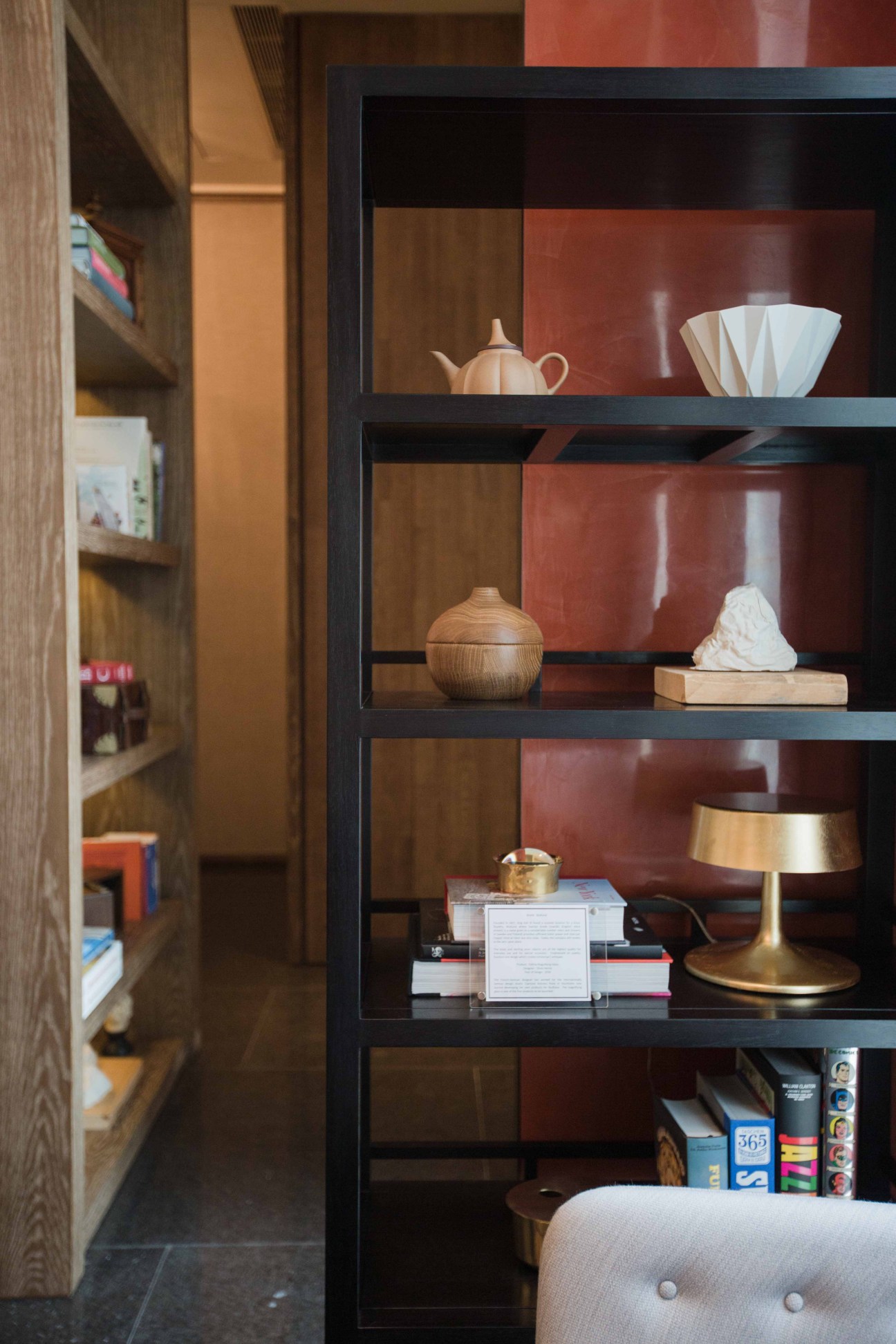 Black shelving unit displaying curated objects including pottery, wooden vessels, books and a brass lamp against a red wall.