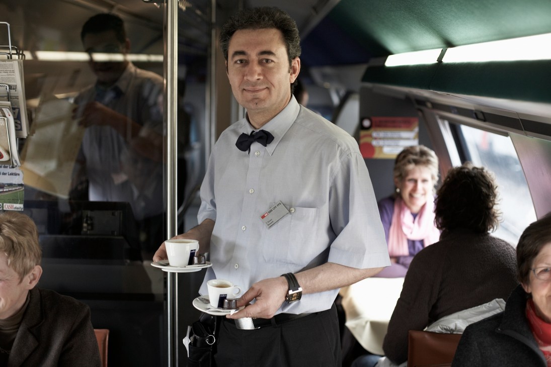 SBB train attendant in white shirt and bow tie serving coffee to passengers onboard.