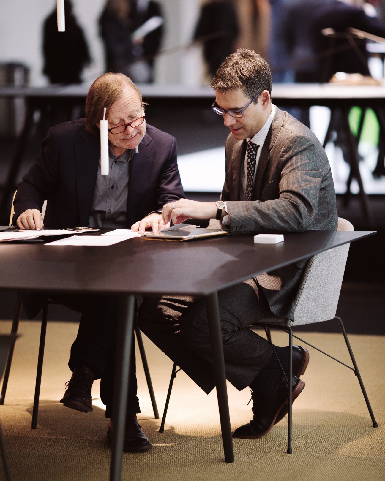 Two businessmen in suits review documents at a table during IMM Cologne furniture fair.