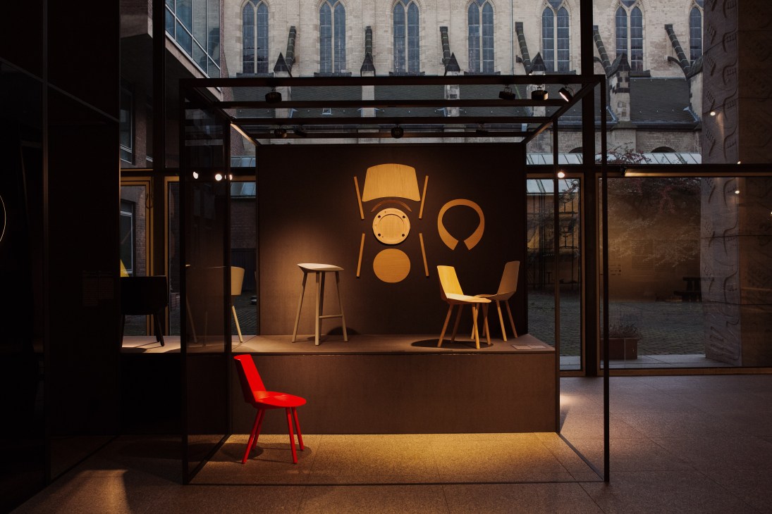 Modern furniture showroom with wooden chairs, stools, and graphic wall display against a dark interior backdrop.
