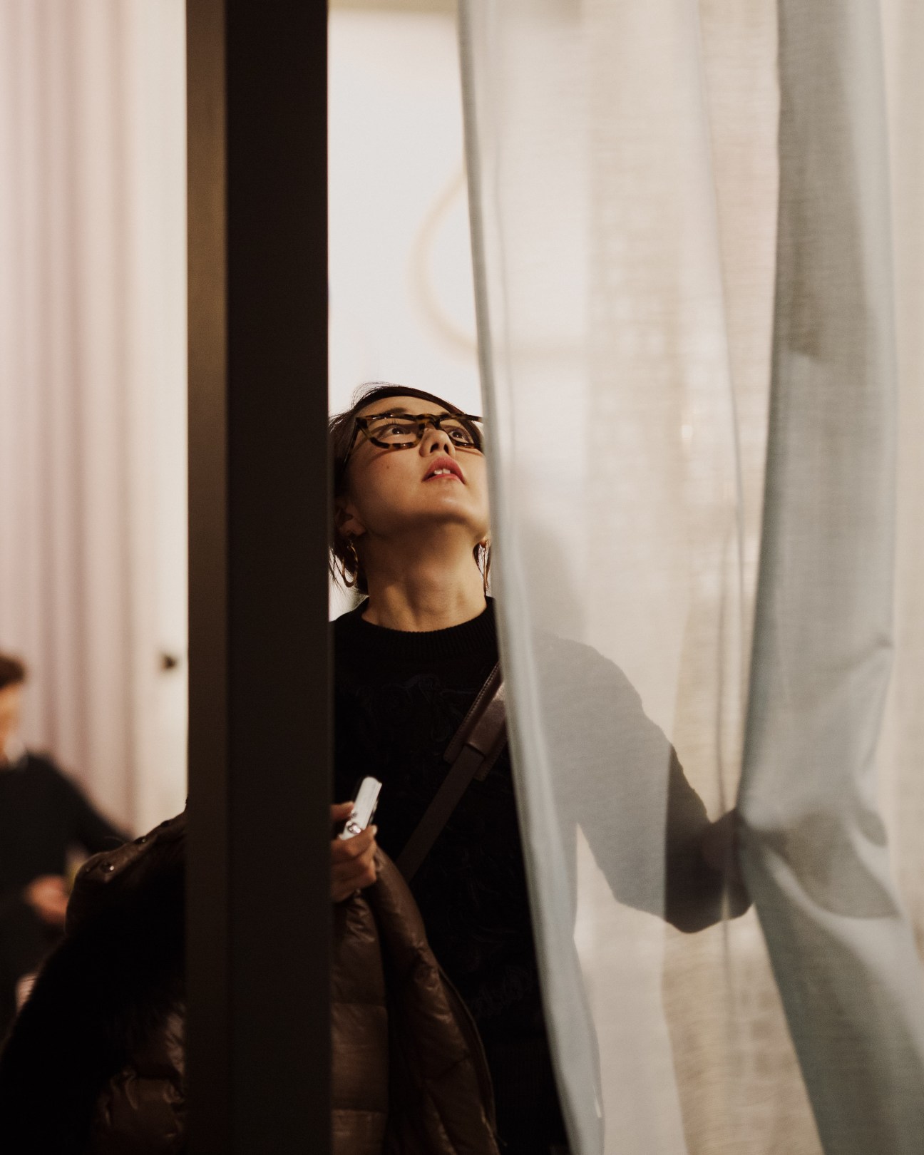A visitor wearing glasses and dark clothing examines hanging white curtains at IMM Cologne furniture fair.