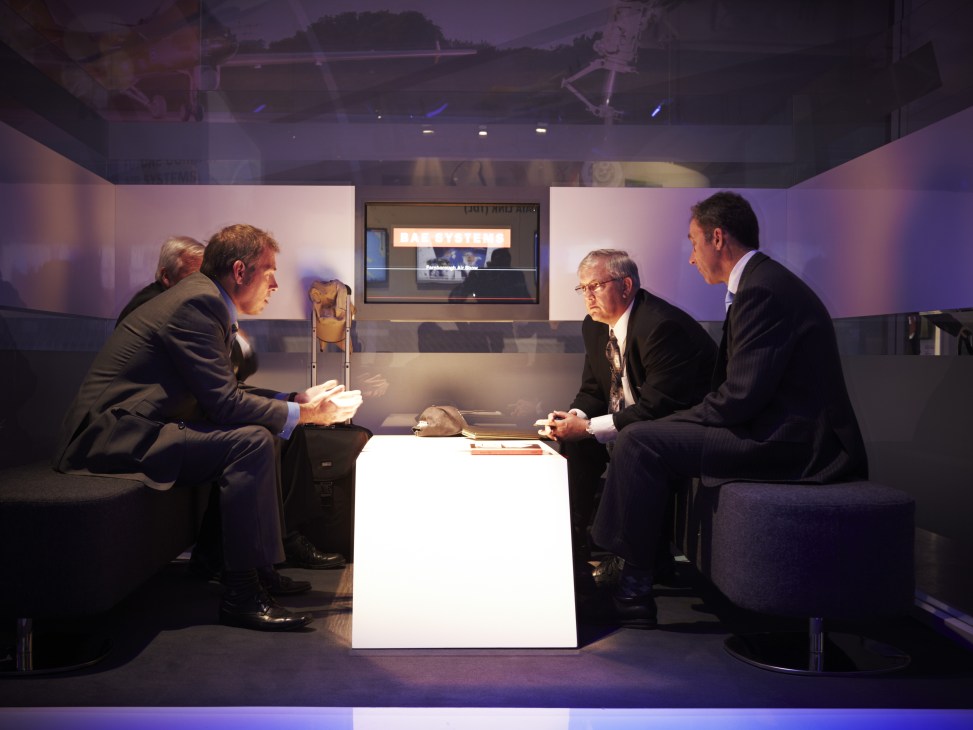 Four businessmen in suits meeting around a white illuminated table at the BAE Systems pavilion.