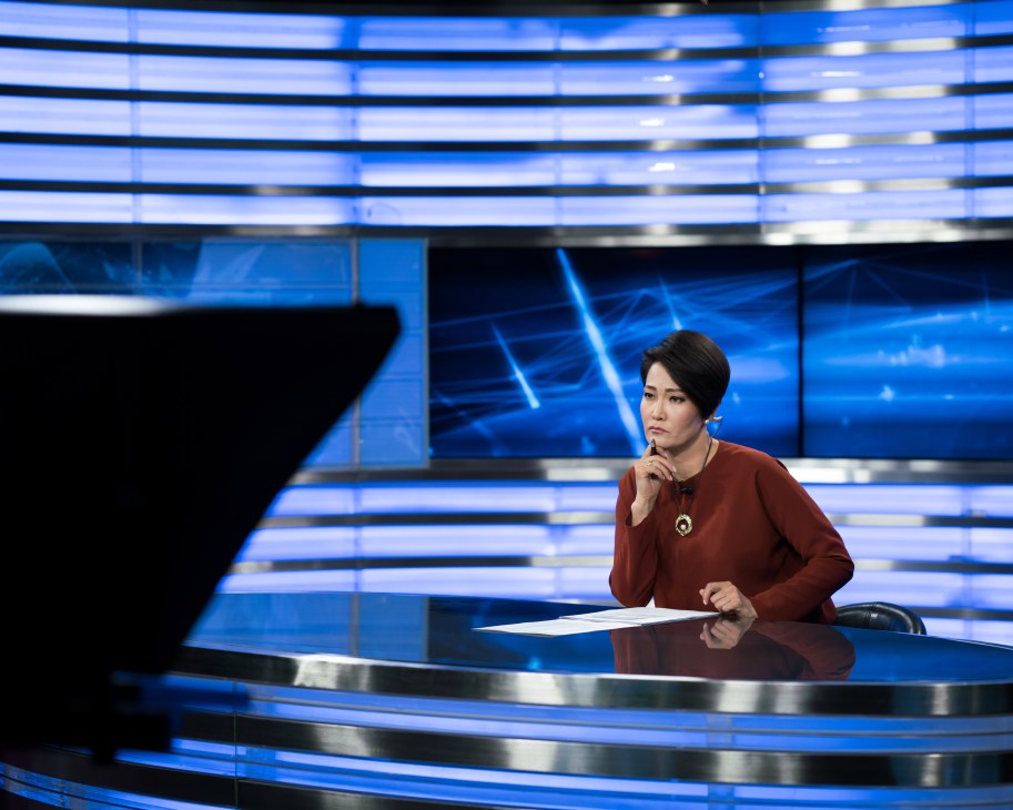 Female news anchor Chimgee presenting Mongol TV news broadcast at desk with blue digital backdrop.