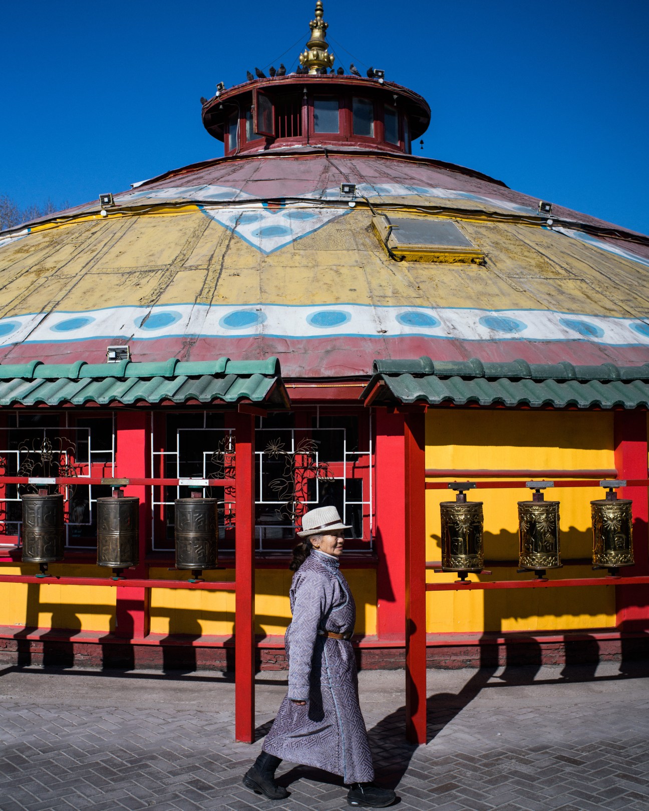 Person in traditional Mongolian del coat walking past Dashichoilin Buddhist monastery with colorful red, yellow and gold exterior.