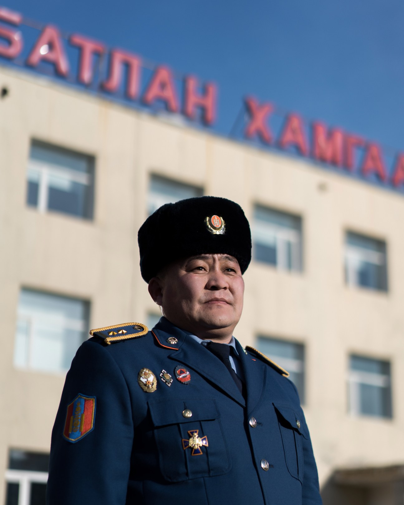 Mongolian military officer in blue uniform with medals standing in front of a building with Cyrillic signage.