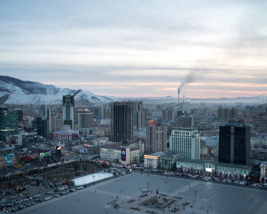 Aerial view of Ulaanbaatar's cityscape with snow-capped mountains in the background and industrial smokestacks visible on the horizon.