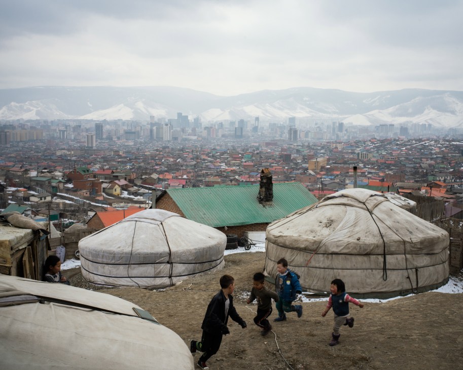 Children play between traditional gers in Ulaanbaatar's ger district with the city skyline in the background.