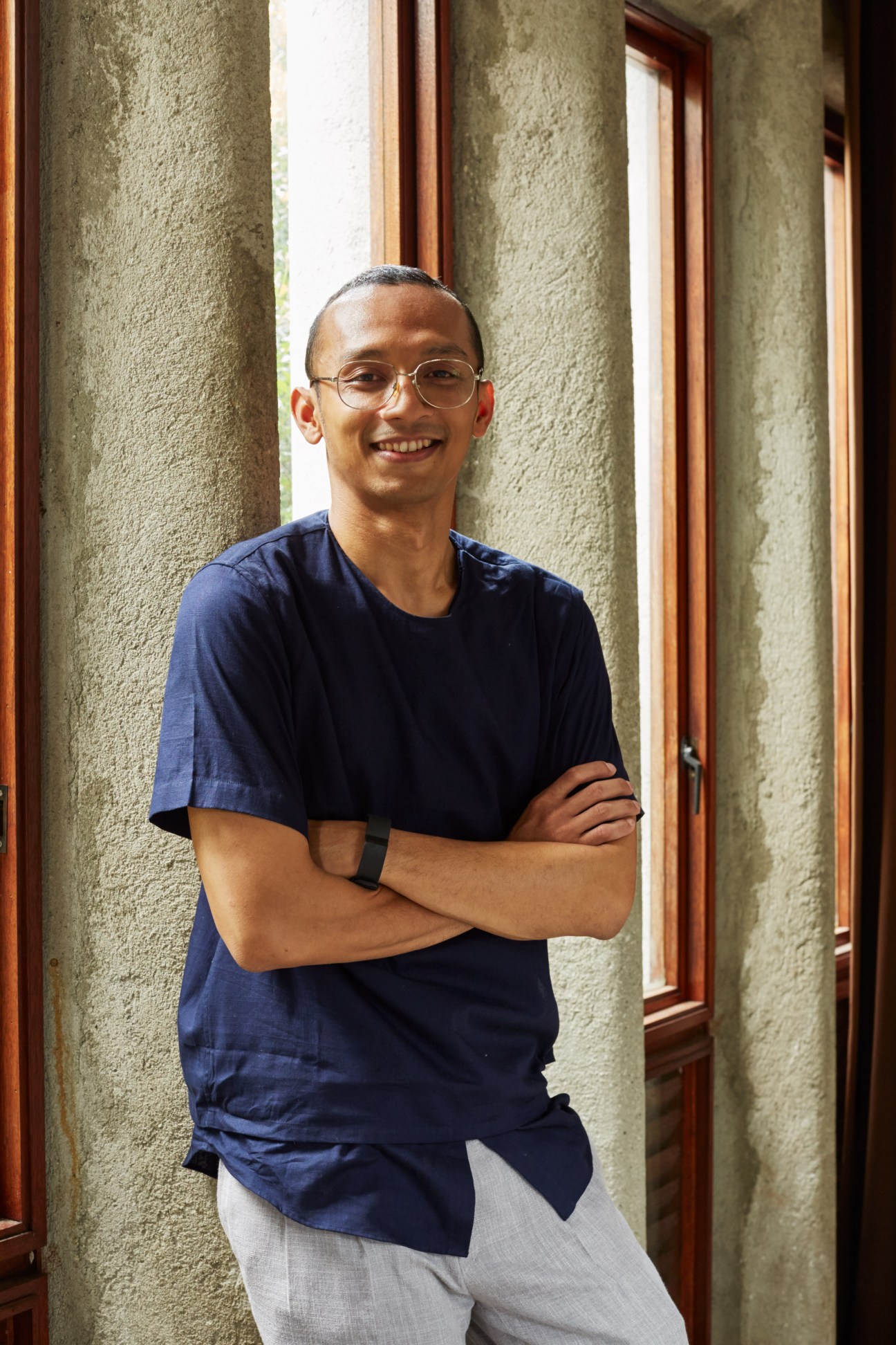 Danny Wicaksono wearing glasses and navy t-shirt with arms crossed, standing by concrete columns and windows