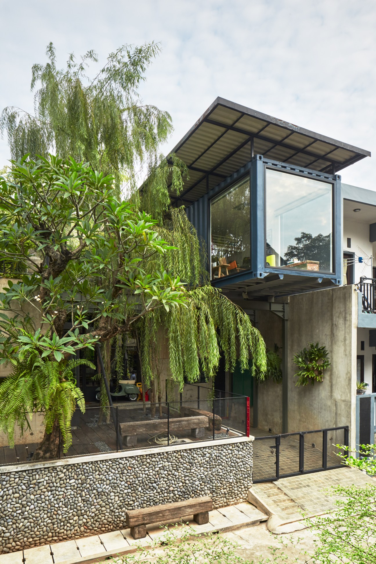 Modern house with glass-walled shipping container extension elevated above concrete base, surrounded by lush tropical plants