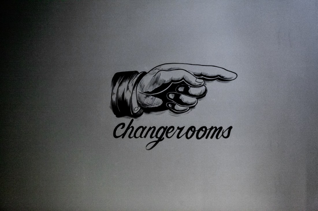 Hand-painted sign featuring a pointing hand illustration above cursive text reading 'changerooms'.