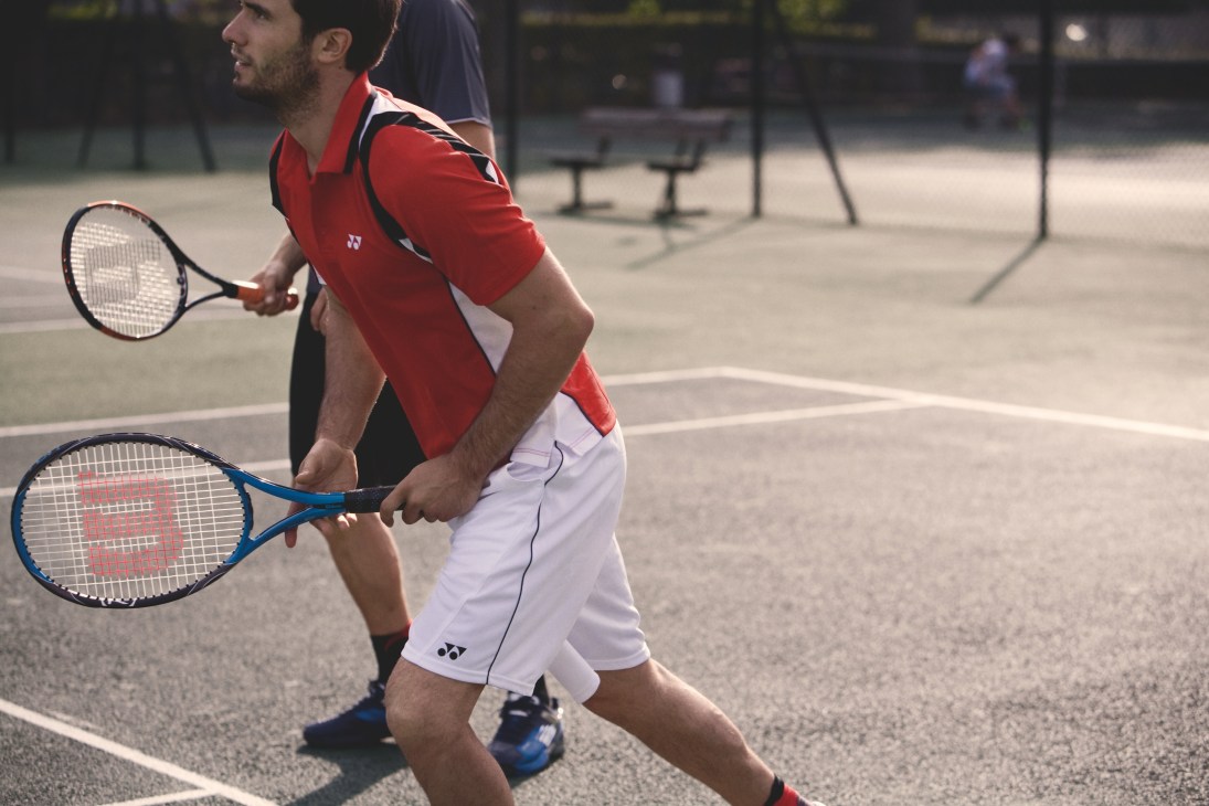 Male tennis player in red polo shirt and white shorts holding Wilson racquet on outdoor court.