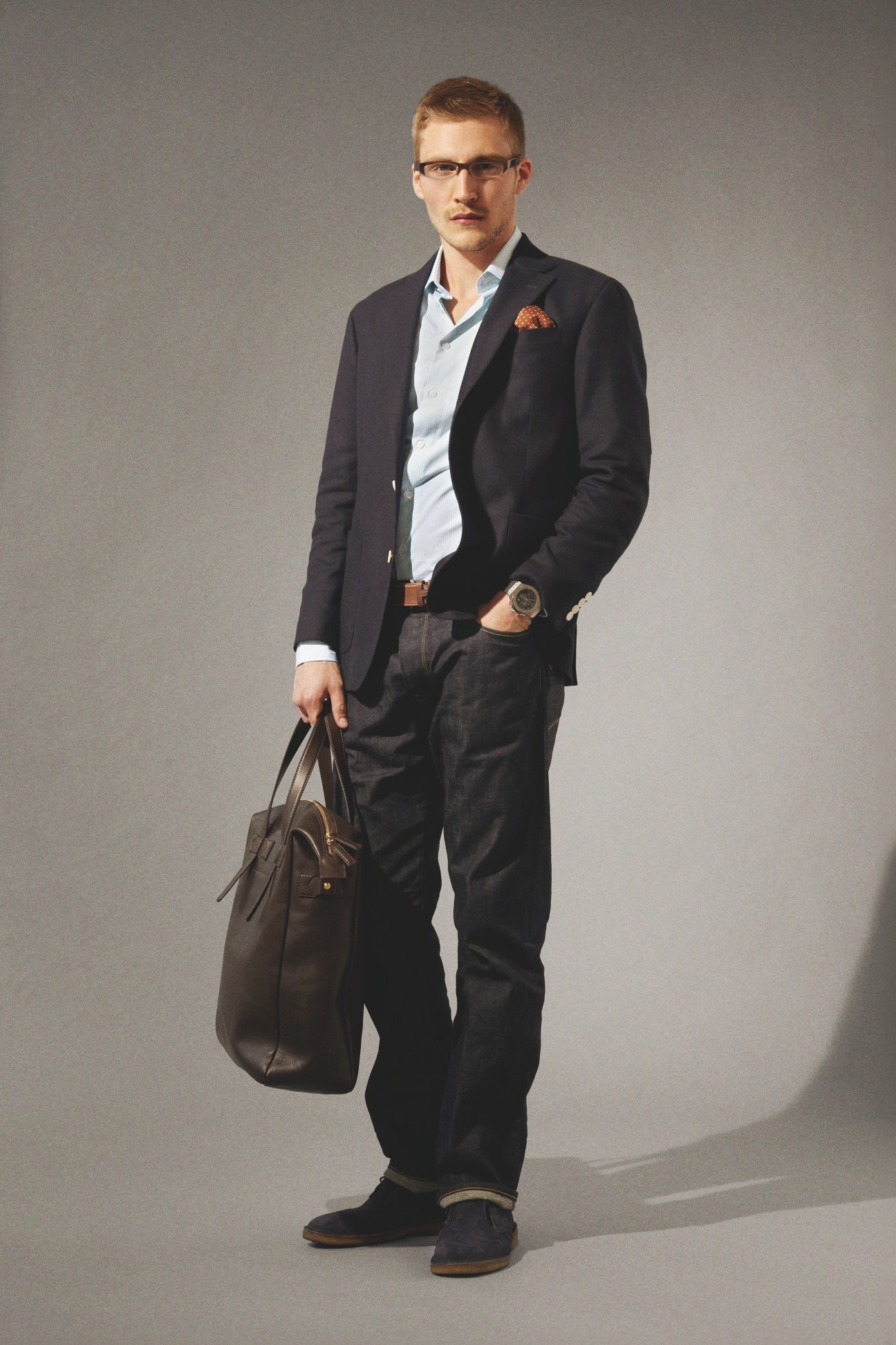 Man wearing black blazer, light blue shirt, dark jeans, brown leather belt and shoes, holding brown leather bag.