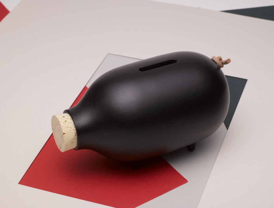 Black ceramic piggy bank with cork nose and wooden tail on geometric red and gray background.