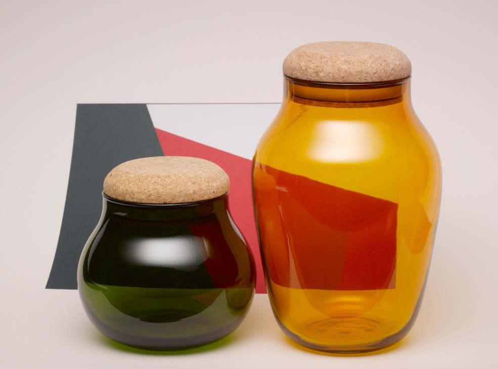 Two glass jars with cork lids containing colored liquids against a geometric background.