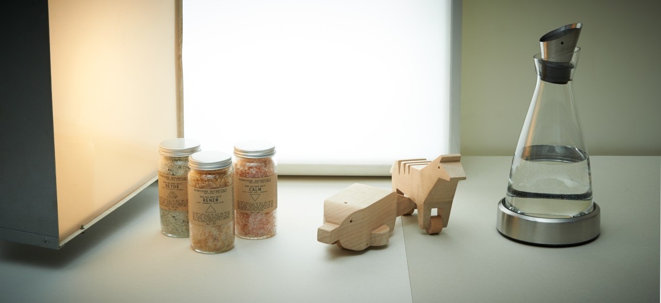 Herbivore Botanicals bath salts in glass jars, wooden animal toys, and Flow cooling carafe on white surface.