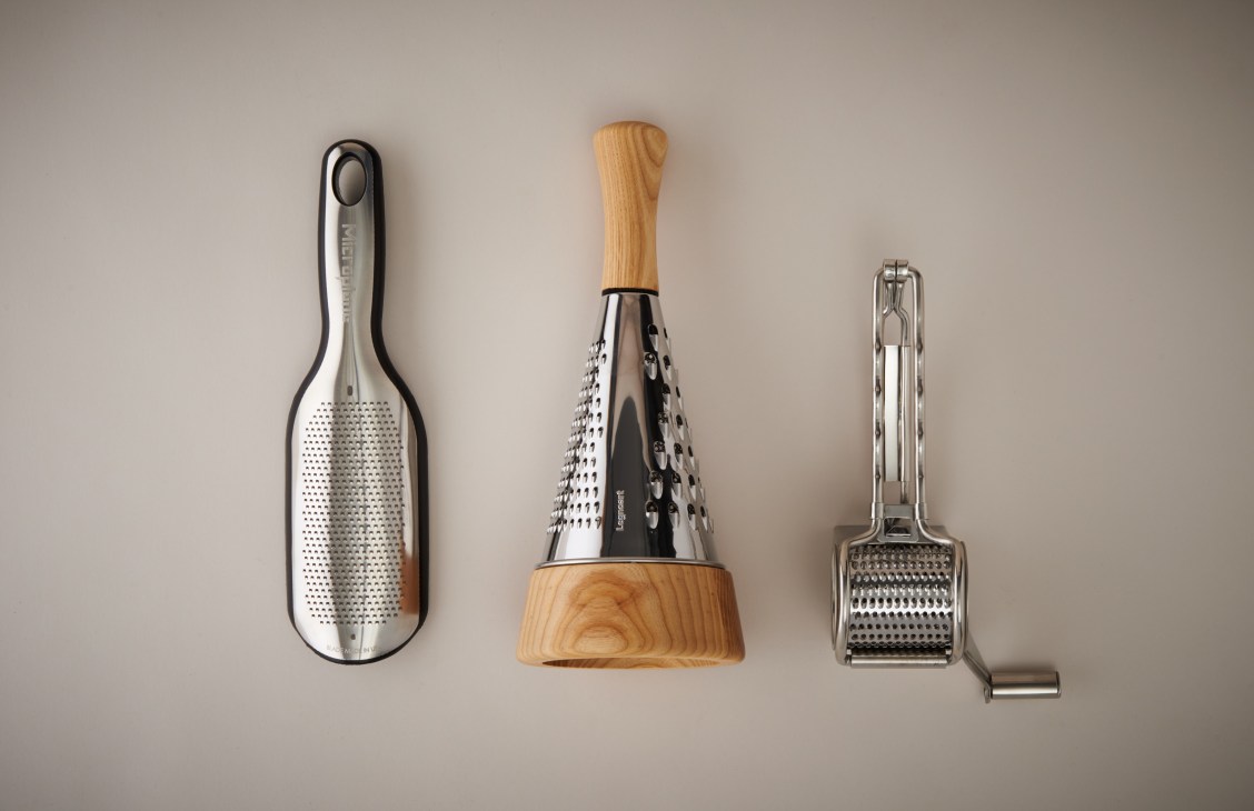 Three cheese graters displayed against a neutral background: a paddle grater with black handle, a cone grater with wooden base, and a box grater with metal frame.