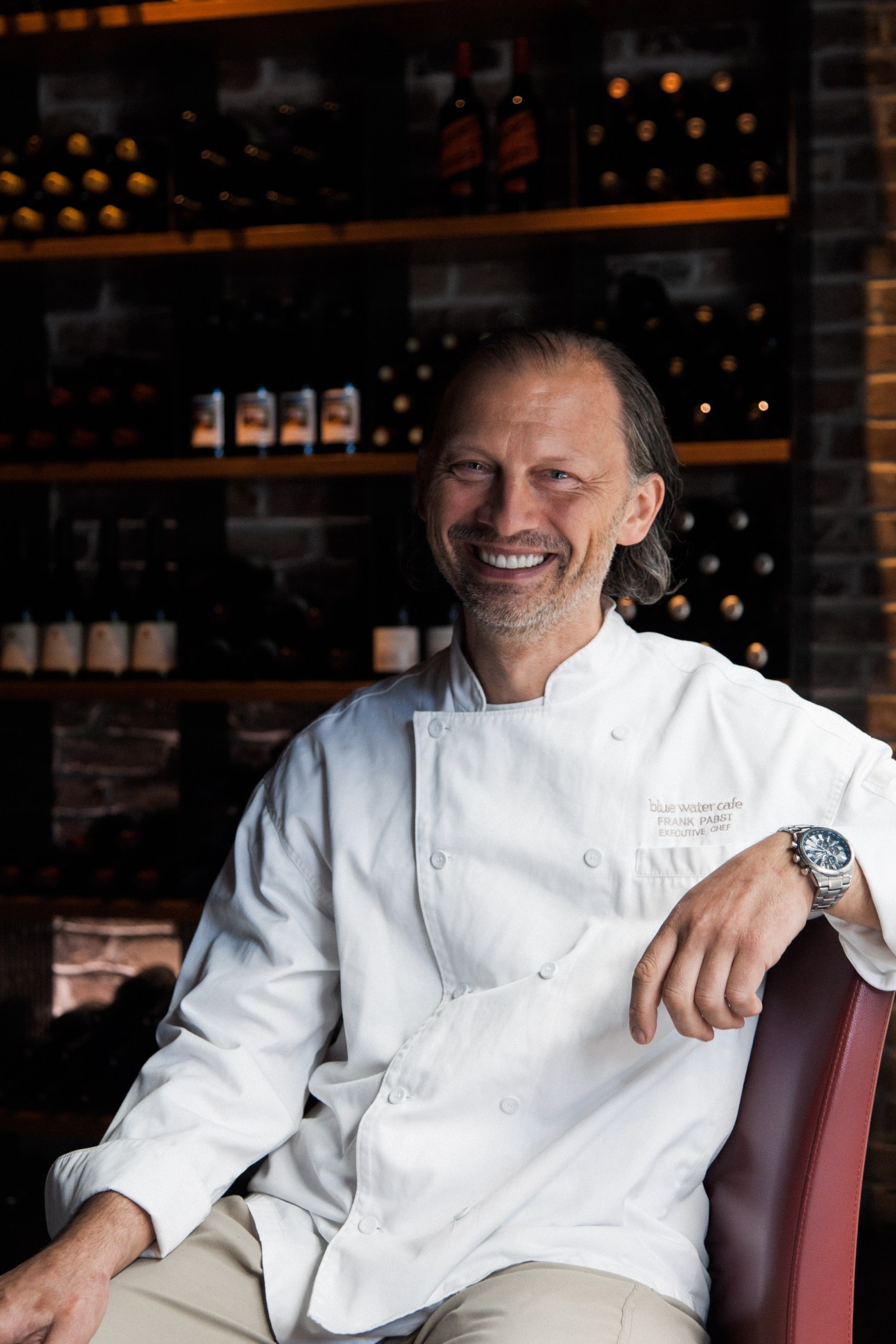 Chef Frank Pabst smiling at Blue Water Café with wine bottles displayed on shelves behind him.