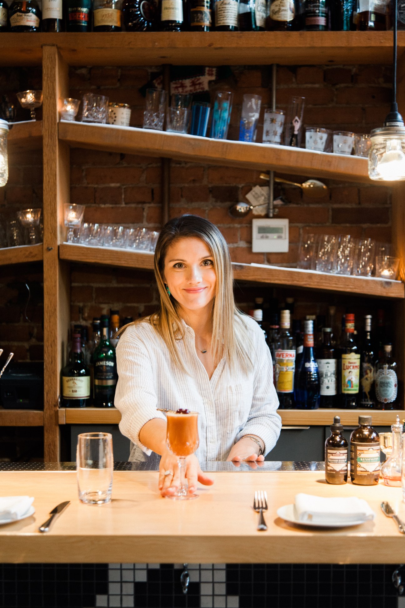 Bartender presenting a cocktail at L'Abattoir's bar with shelves of spirits behind.