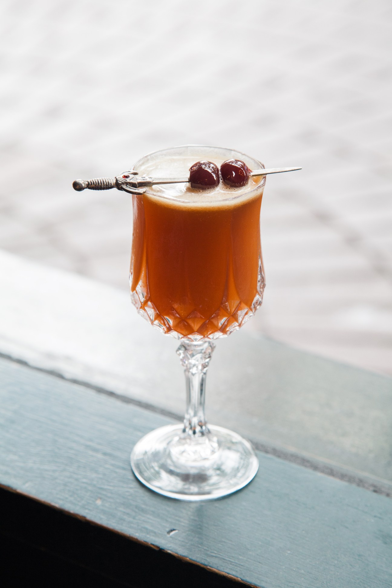 Amber cocktail with two dark cherries on a pick, served in a coupe glass with ocean backdrop.