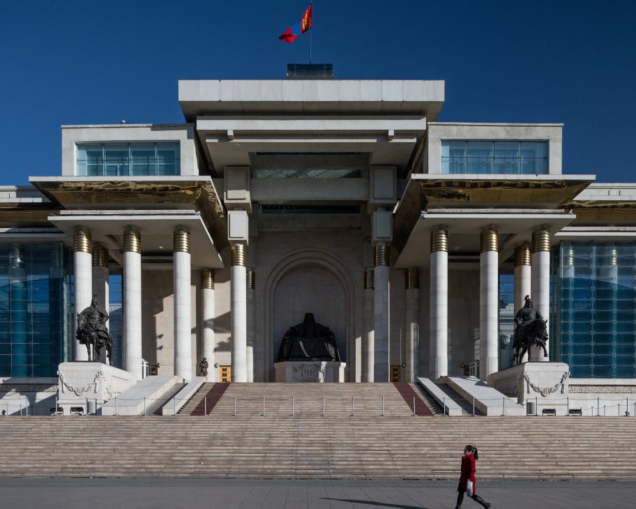A modernist government building with white columns, bronze capitals, and a Mongolian flag atop under clear blue sky.
