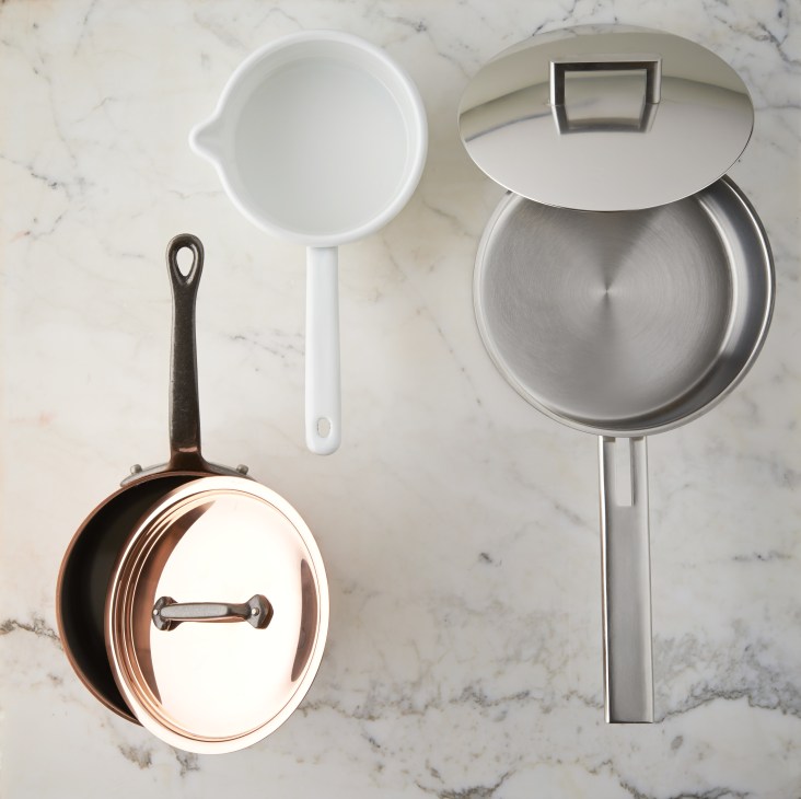 Three saucepans displayed on marble surface: copper pan with lid, white handled pan, and stainless steel pan with lid.