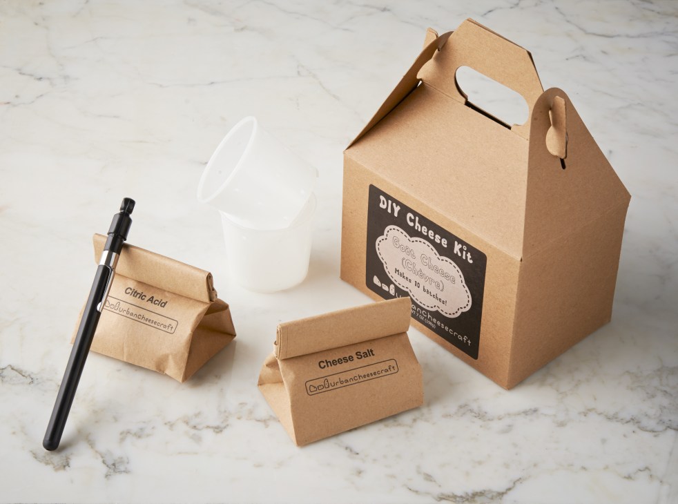 Urban Cheesecraft DIY cheese-making kit with cardboard box, ingredient packets, and black applicator on marble surface.