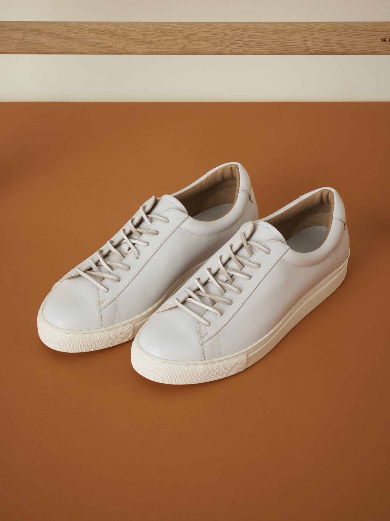 Pair of white leather lace-up sneakers with cream-colored soles on a tan background.
