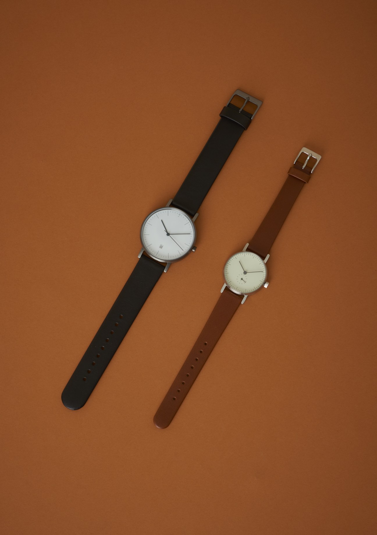 Two Stock Watches unisex timepieces with white dials and minimalist design on black and brown leather straps against a tan background.
