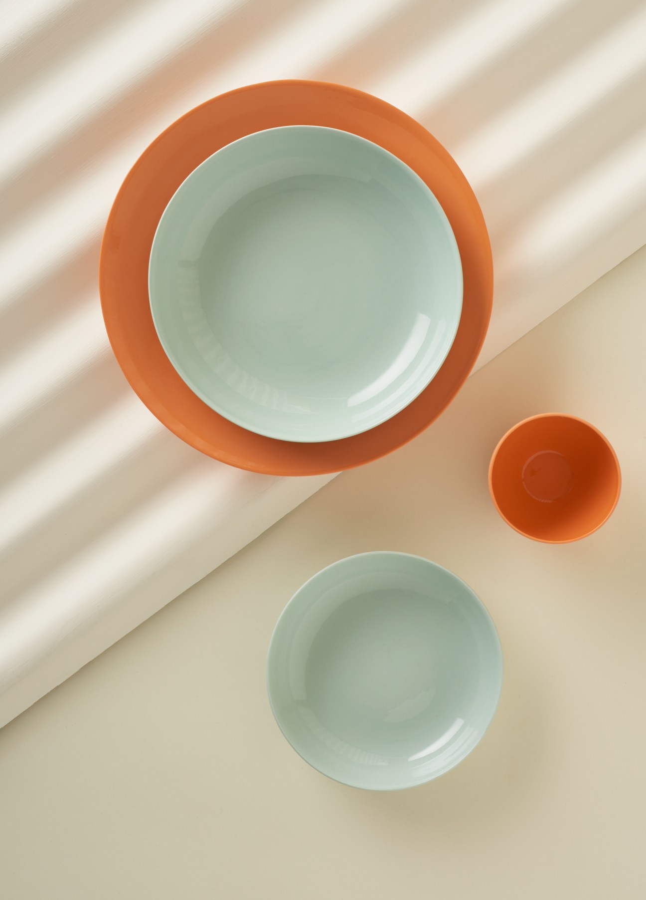 Mud Australia ceramic bowls in pastel mint green and terracotta orange arranged on a beige surface with diagonal shadow.