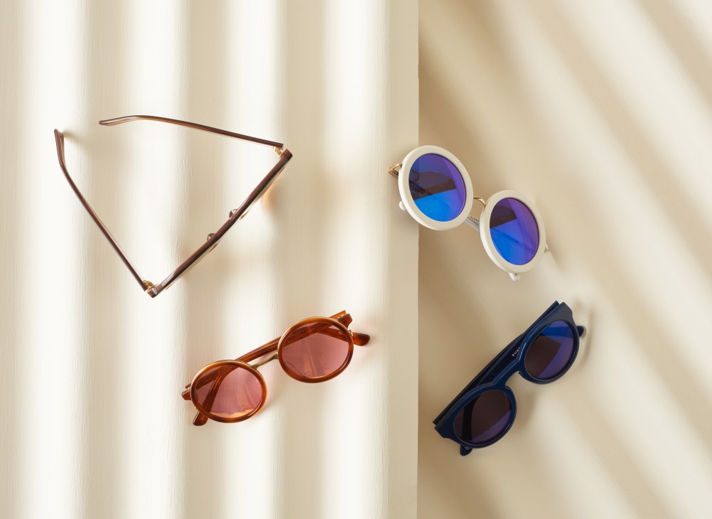 Four pairs of Sunday Somewhere sunglasses with various frame styles and colored lenses arranged on a neutral surface.