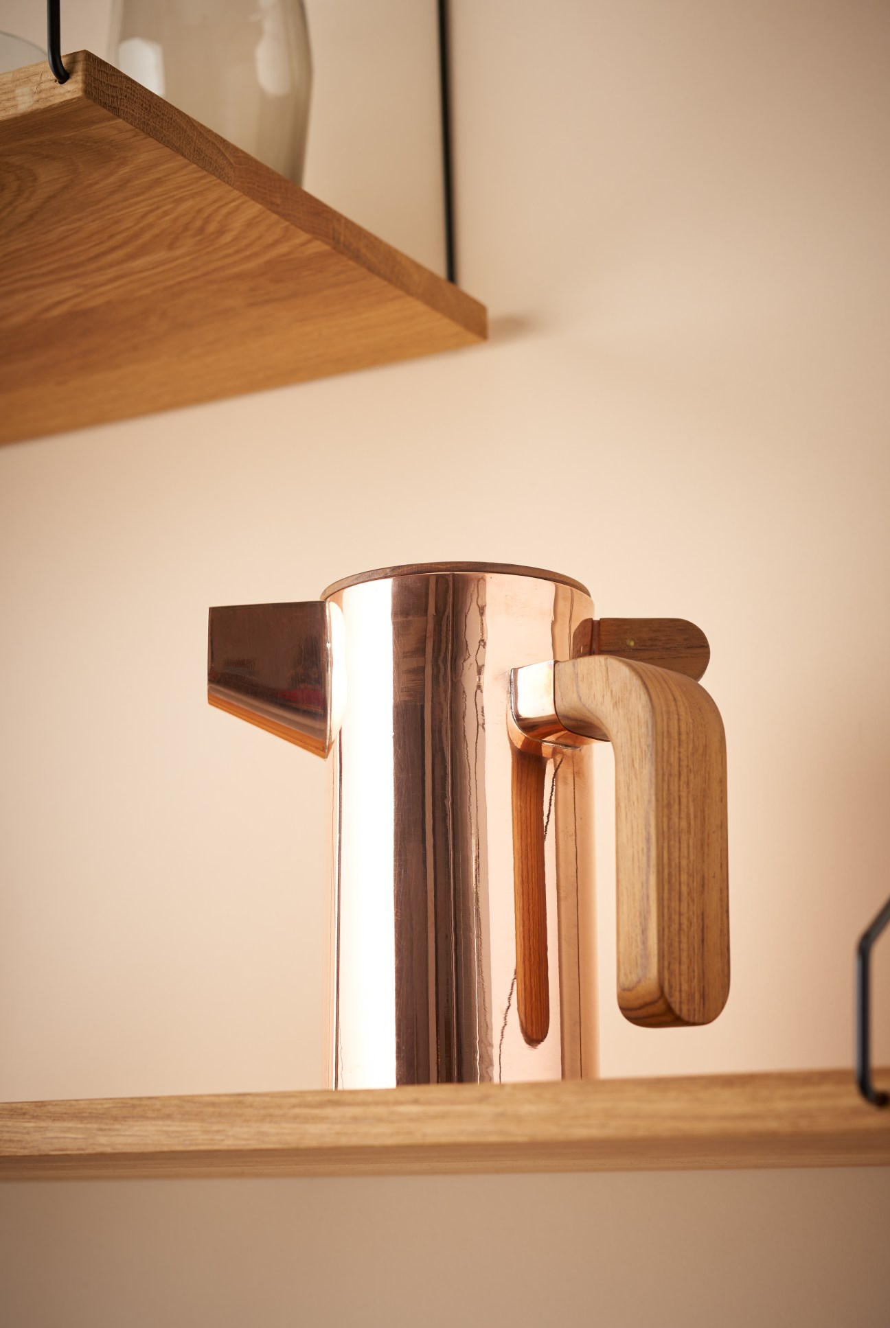 A polished copper coffee pot with wooden handle by Breaktime Kit on a wooden shelf.