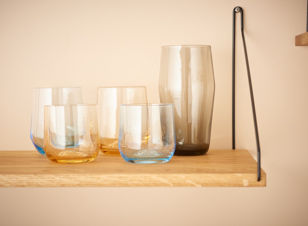Handblown soda-lime glasses in yellow, grey and blue hues by Mazama Wares displayed on a wooden shelf.