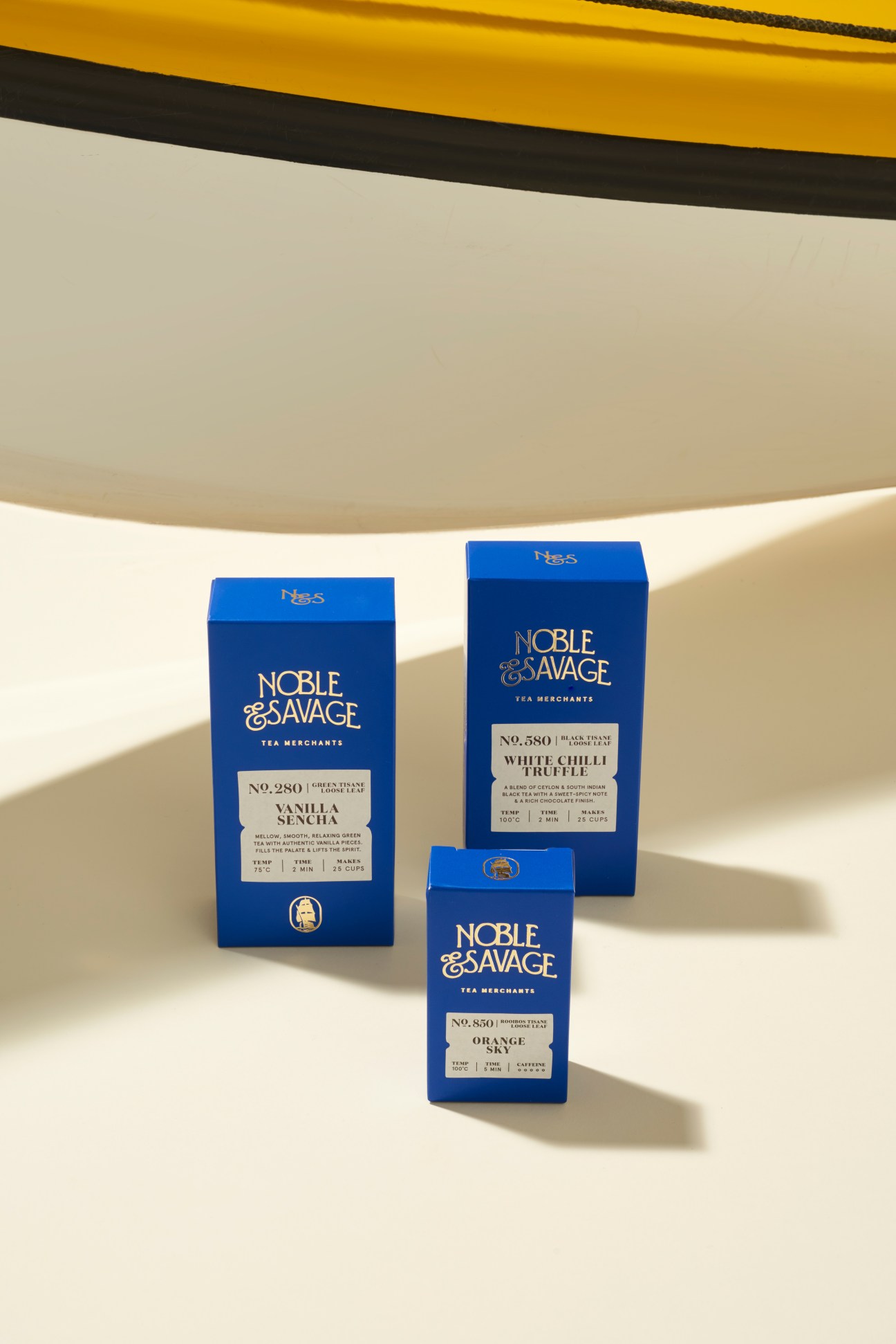 Three blue Noble & Savage tea boxes displayed on a white surface with dramatic shadows and yellow curved element above.