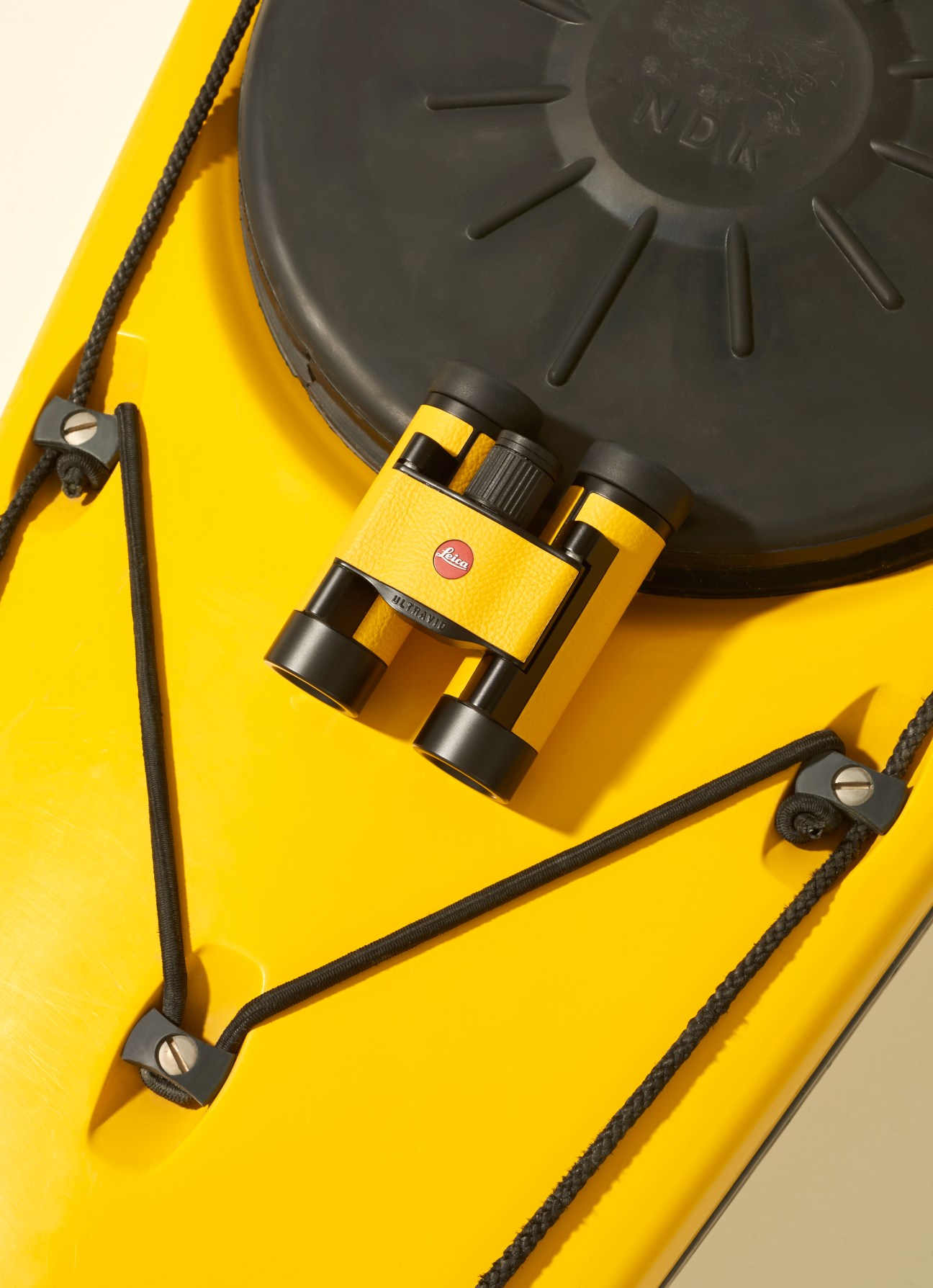 Yellow Leica binoculars with black straps resting on a yellow surface with a dark lens visible.