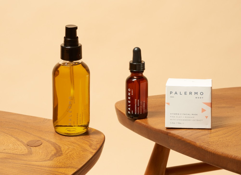 Palermo Body skincare products including a golden oil spray, red serum dropper bottle, and white Vitamin C facial mask box displayed on wooden surfaces.