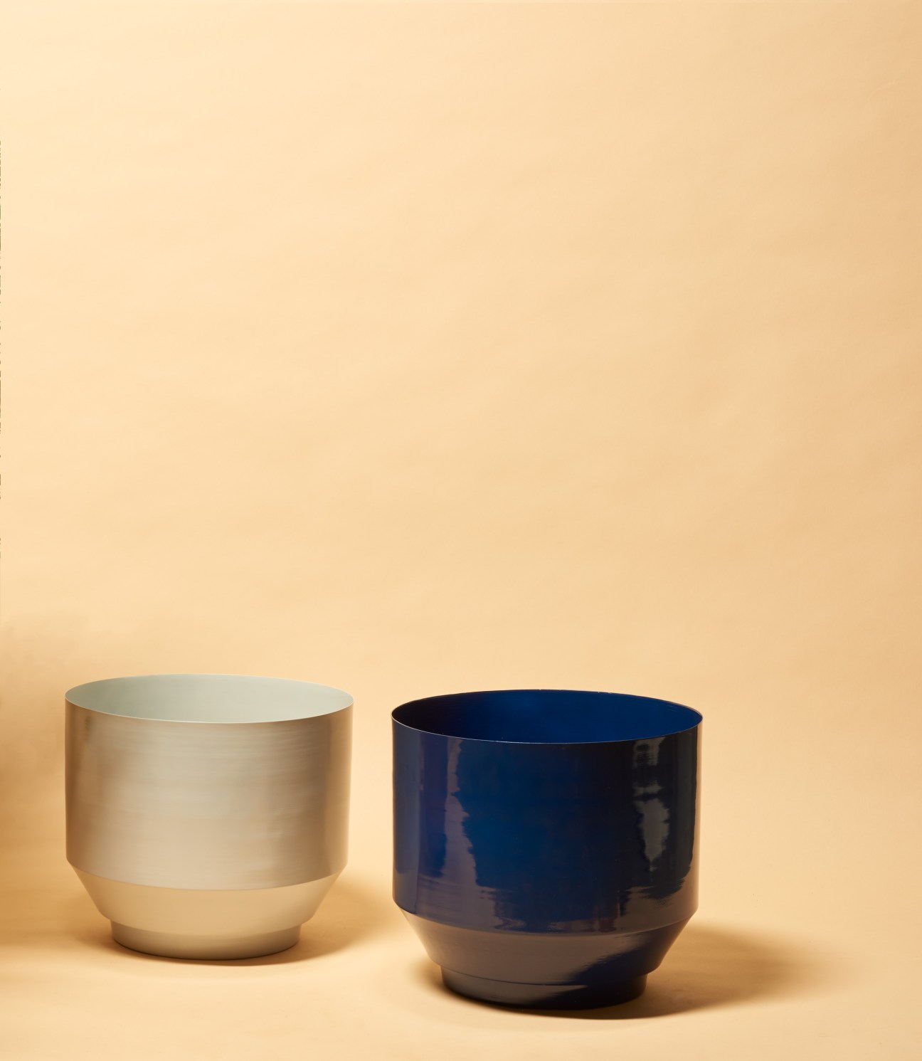 Two ceramic planters by Yield against a beige background: one cream-colored and one dark blue with metallic bases.